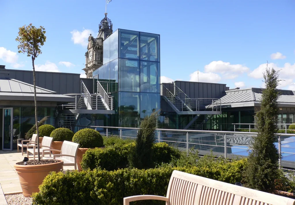A sunny roof terrace with potted plants and wooden benches in front of a modern glass elevator structure.