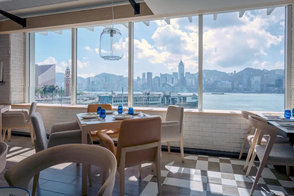 Restaurant interior with table set-up near floor-to-ceiling windows providing panoramic harbor views, under a bright, clear sky.