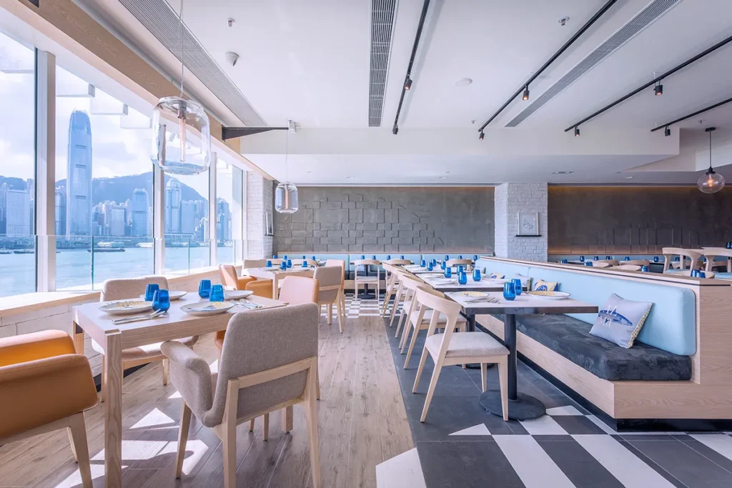 Modern restaurant interior with a mix of booth and table seating, geometric floor design, against a backdrop of a city skyline view.