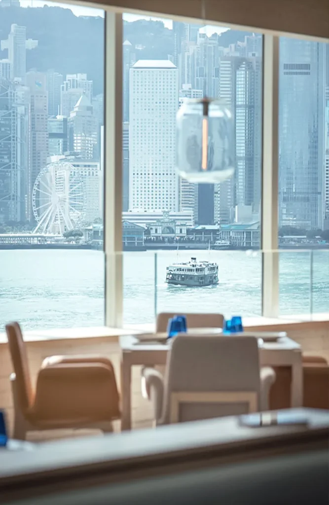 Elegant restaurant dining area with large windows overlooking Hong Kong harbor and a ferry.