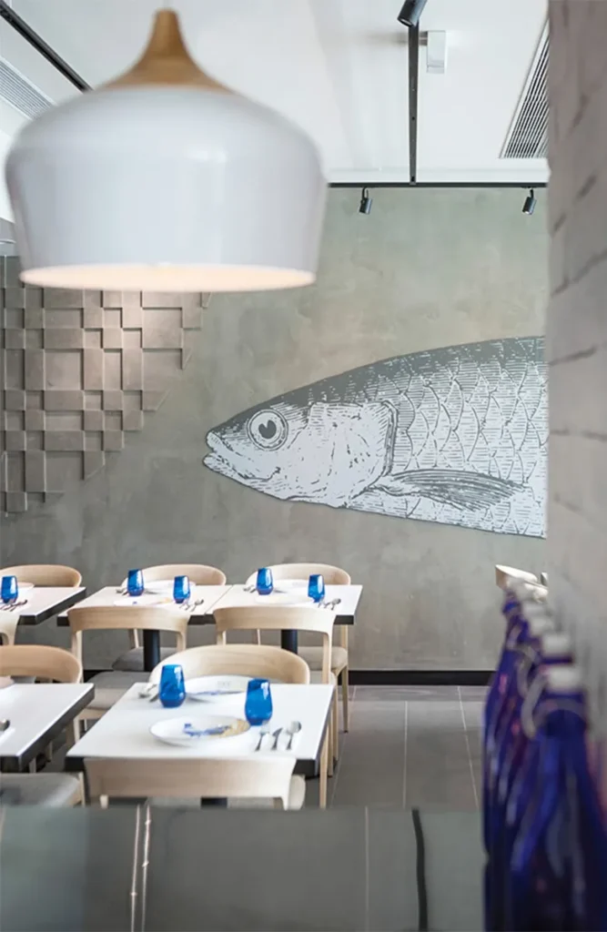 Modern restaurant interior with a large fish illustration on the wall, geometric hanging lights, and blue glassware on light wood tables.