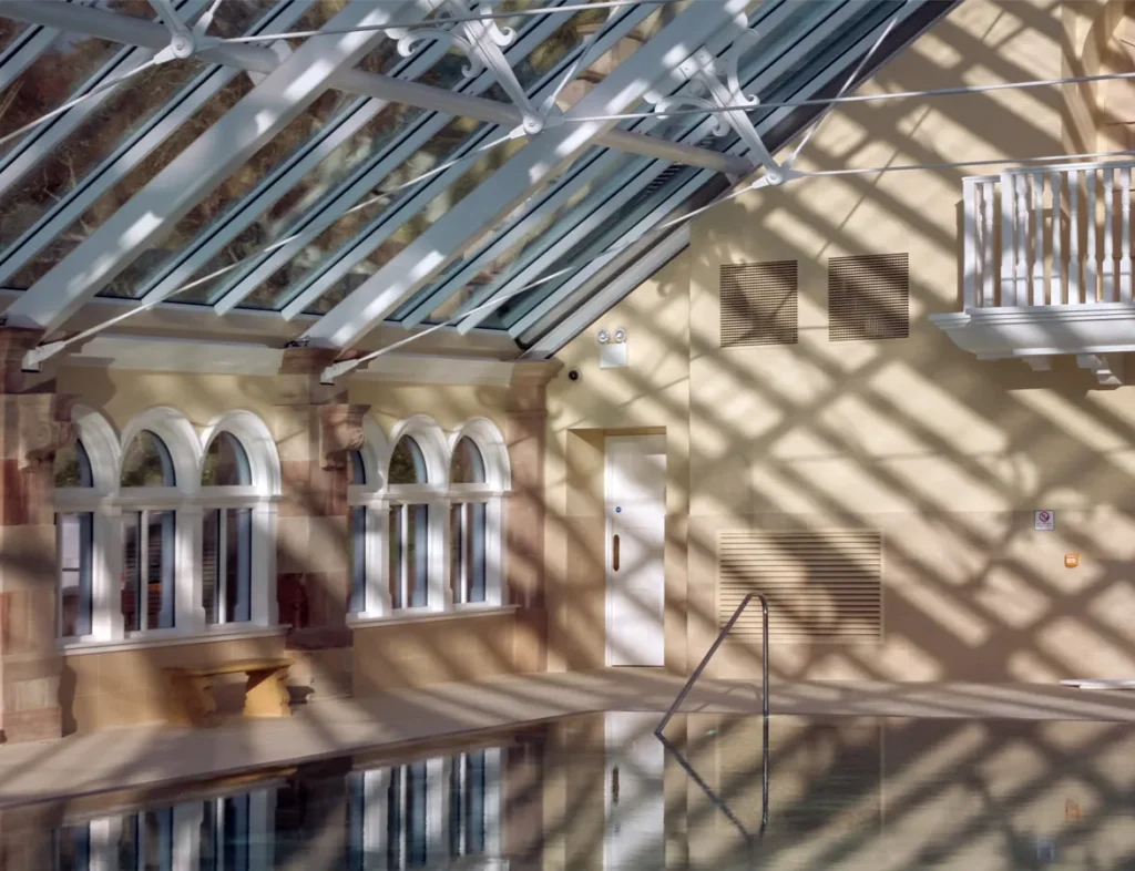 Sunlit indoor pool casting shadows on the stone wall and glass ceiling.