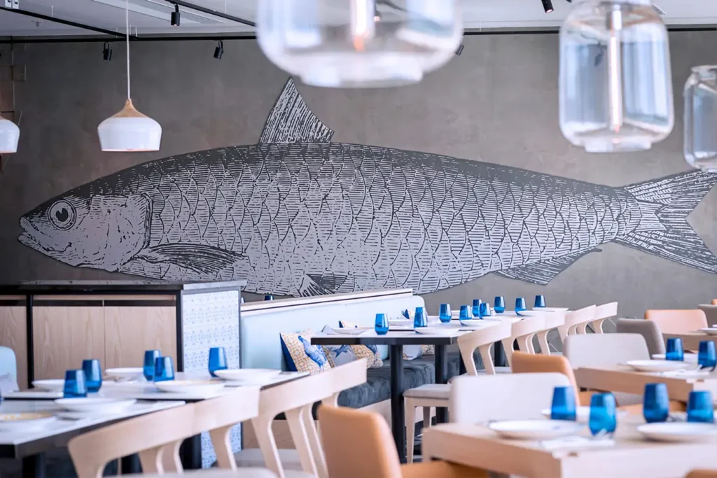Dining room in a seafood restaurant with a large fish mural, hanging glass lights, and blue accents.
