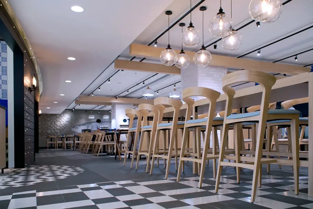 A contemporary restaurant interior featuring light wooden high stools at a high table, a checkerboard floor, pendant lights, and blue patterned accents on the wall.