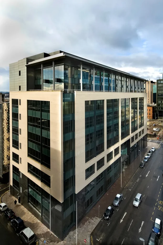 Elevated view of a contemporary office building at a street corner, showcasing a juxtaposition of reflective glass and neutral stone facades under a dynamic sky.